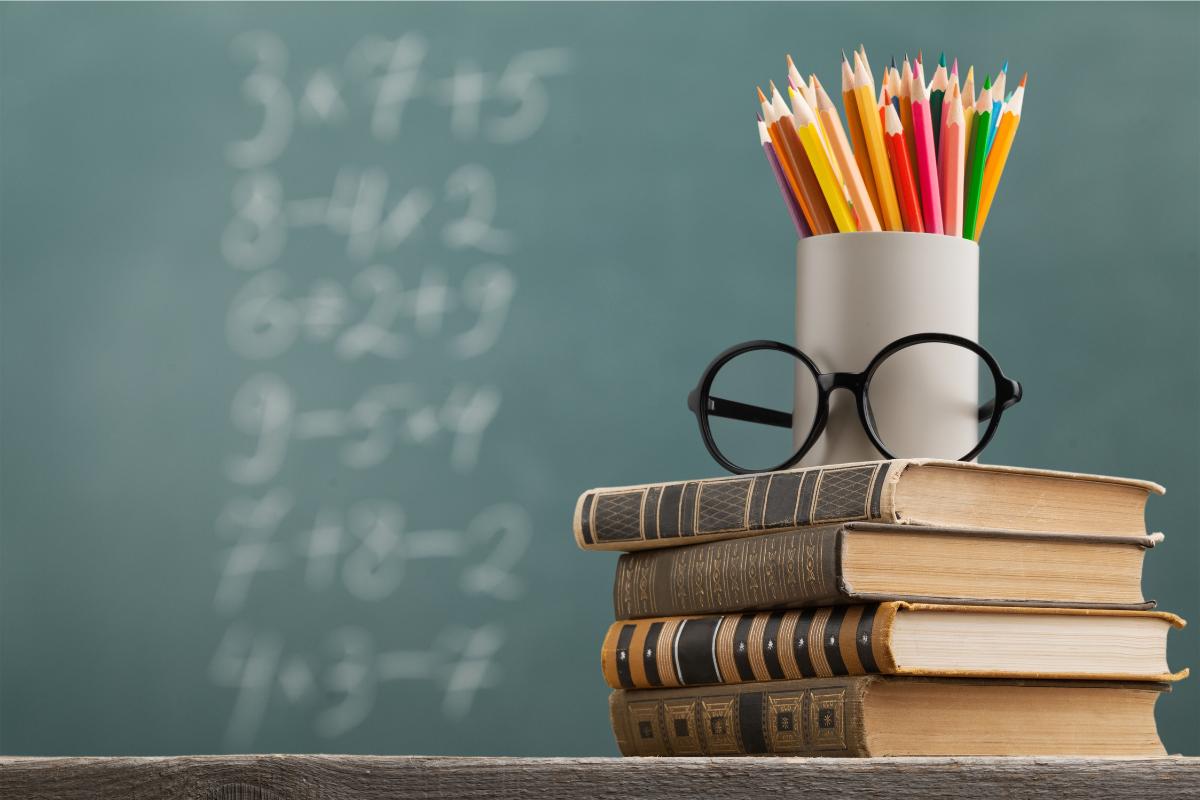 libri su cattedra con porta matite pieno e occhiali, sullo sfondo la lavagna con numeri